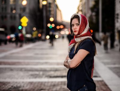 A person wearing a red and white headscarf is turned to the side with their arms crossed, but is looking directly into the camera